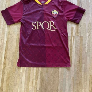 AS Roma vinröd fotbollströja SPQR - Snygg vinröd AS Roma fotbollströja med gul krage och klubbmärke på bröstet. Framsidan har diskret mönster och SPQR-tryck i guld. Tillverkad i lätt och ventilerande material, perfekt för match eller träning.