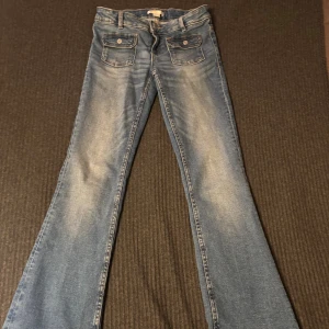 Blå bootcut jeans från Gina Tricot - Snygga blå bootcut jeans från Gina Tricot med slitningar och två fickor med lock framtill. Klassisk femficksmodell med bälteshällor och knappstängning. Perfekta för dig som gillar en retro vibe och vill ha ett par jeans med lite utsvängda ben.