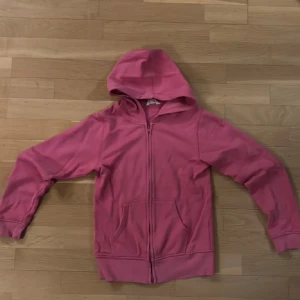 Rosa hoodie med dragkedja och huva - Säljer en rosa hoodie med dragkedja framtill och huva. Tröjan har två fickor på magen och ribbade muddar vid ärmslut och nederkant. Perfekt för chill dagar eller när du vill vara extra comfy.