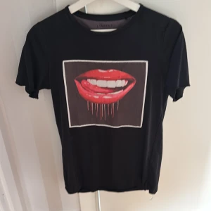 Svart t-shirt med läppar från Limitato - Svart t-shirt från Limitato med korta ärmar och en stor grafisk print framtill av röda läppar med vita tänder. T-shirten har en klassisk rund halsringning och är gjord i mjukt material som känns skönt mot huden. Perfekt för dig som vill sticka ut med en cool detalj.