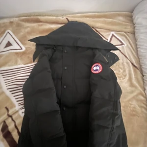 Svart dunjacka från Canada Goose S - Svart dunjacka från Canada Goose med huva och klassisk logga på ärmen. Jackan är quiltad och har knappar framtill. Perfekt för kalla dagar och har en clean, stilren look som funkar till det mesta. DUSTBAG/PÄLS INGÅR
