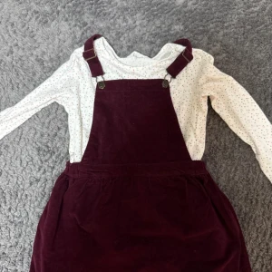 Vinröd manchesterklänning med hängslen - Supersnygg vinröd kortklänning i mjuk manchester med klassiska hängslen och metallspännen. Klänningen har en rak passform och är perfekt att styla med en långärmad topp under. Enkel och cool look som funkar året runt.