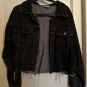 Svart croppad jeansjacka - Cool svart jeansjacka med croppad passform och råa, fransiga kanter nertill. Klassisk krage, knäppning framtill och två bröstfickor med lock.