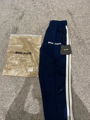 Palm Angels blå trackpants med vita ränder - Palm Angels trackpants i mörkblått med vita sidoremsor och svart tunn linje. Byxorna har elastisk midja med snörning och logotypen broderad i vitt framtill. Snygg streetwear-vibe och skön passform, perfekt för chill eller sportig stil. Byxorna är i bra skick förutom att det finns 2st brännmärken som man kan se på 3:e bilden. Nypris: 5499kr, mitt pris 799kr. 
