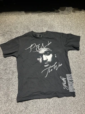 Pop Smoke x VLONE svart t-shirt - Svart t-shirt med Pop Smoke-tryck och texten 'The Woo' på framsidan, samt stor vit rökformad V på ryggen. Långa låtlistor tryckta nertill. Klassisk passform och rund hals. Perfekt för dig som gillar streetwear och hiphop.