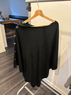 Svart bolerotopp med knappar - Svart bolerotopp med bred halsringning och dekorativa silverfärgade knappar längs ena axeln och ärmen. Toppens material är mjukt och har ett lätt fall, vilket ger en chill och avslappnad vibe. Perfekt för dig som gillar minimalistisk stil med en twist.