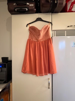Korallrosa axelbandslös spetsklänning - Superfin korallrosa kortklänning med axelbandslös design och hjärtformad byst. Överdelen är täckt av spets och kjolen är lätt och luftig med vidd. Perfekt för dig som vill sticka ut med en romantisk och feminin look. Storlek M. Helt ny med tags/ prislapp fortfarande kvar. 
