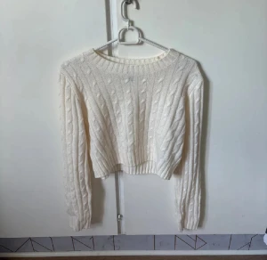 Vit stickad croppad tröja från H&M Divided - Säljer en vit stickad tröja från H&M Divided i storlek M. Tröjan är croppad med kabelstickat mönster på framsidan, ribbad hals och muddar samt långa ärmar. Perfekt för dig som gillar en clean och chill stil.