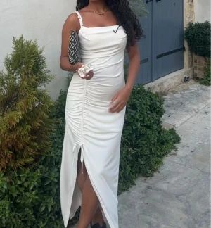 Vit långklänning  - Säljer en elegant vit långklänning med smala axelband och snygg snörning framtill som ger en  rynkad effekt. Klänningen har en slits framtill och sitter figurnära. Använd 2 gånger så som ny!