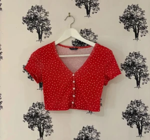 Röd croppad topp med hjärtan  - Säljer en röd croppad topp från Shein med små vita hjärtan över hela tyget. Toppen har v-ringning, korta ärmar och knappar framtill. Perfekt för dig som gillar söta detaljer och vill ha en chill och trendig look.