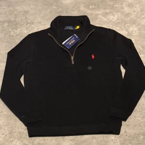 Svart half zip tröja Polo Ralph Lauren - Svart långärmad tröja från Polo Ralph Lauren med half zip och ståkrage. Tröjan har det klassiska röda Polo-logot broderat på bröstet och är tillverkad i mjuk bomull. Perfekt för dig som gillar stilrena och tidlösa plagg med snygga detaljer. Personligen tycker jag att den passar mer som s 
