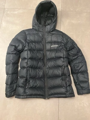 Svart dunjacka Peak Performance - Svart dunjacka från Peak Performance med huva och dragkedja framtill. Jackan har ett quiltat mönster och logotyp på bröstet. Perfekt för kalla dagar med sin puffiga design och långa ärmar. Snygg och funktionell för vinter och höst. Storlek är 170/xs