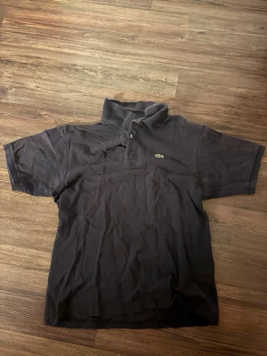 Svart piké från Lacoste - Svart pikétröja från Lacoste med klassisk krage och två knappar framtill. En av knapparna är borta. Den är väldigt använd och krympt en del under åren. Därav priset.
