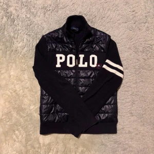 Svart cardigan från Polo Ralph Lauren - Svart kofta från Polo Ralph Lauren med stora vita bokstäver 'POLO' framtill och RL-logga i ett hjärta. Framsidan har quiltade paneler i syntetmaterial och dragkedja. Stickade ärmar med två vita ränder på ena armen och ribbad krage. Perfekt för dig som gillar sportig och stilren look.