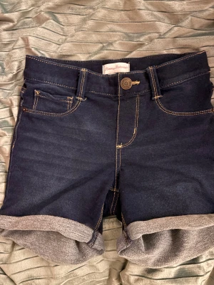 Mörkblå jeansshorts Tommy Bahama - Snygga mörkblå jeansshorts från Tommy Bahama med klassisk femficksdesign, kontrastsömmar och uppvikta benslut. Perfekta för sommardagar och har en normal passform med knapp och dragkedja framtill.