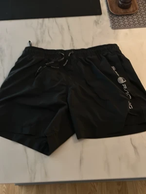 Svarta Sail Racing badbyxor med snörning - Svarta badbyxor från Sail Racing med elastisk midja och snörning. Tillverkade i lätt syntetmaterial med meshfoder för extra komfort. Snygg logga på ena benet och diskret Sail Racing-märke bak. Perfekta för sommarens alla äventyr. Helt oanvända