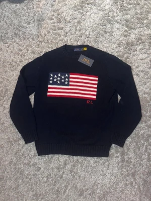 Mörkblå stickad tröja Polo Ralph Lauren - Mörkblå stickad tröja från Polo Ralph Lauren med amerikansk flagga framtill och RL-broderi. Klassisk rund halsringning och ribbade muddar. Perfekt för dig som gillar preppy stil och vill sticka ut med en ikonisk detalj.