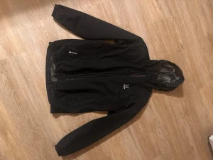 Svart vindjacka från Haglöfs med huva - Svart vindjacka från Haglöfs med GORE-TEX-material och justerbar huva och midja. Jackan har dragkedja framtill, två sidofickor med dragkedja och en stilren logga på bröstet. Perfekt för blåsiga och regniga dagar och outdoor-äventyr.