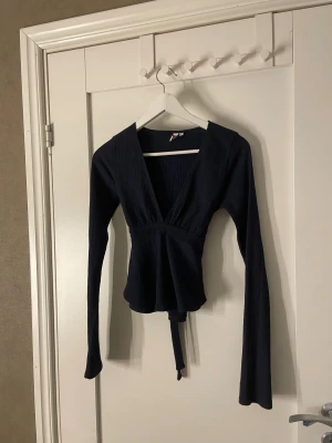Mörkblå omlottopp från Nelly - Säljer en mörkblå omlottopp från Nelly med långa ärmar och knytband i midjan. Toppen har en djup v-ringning. Perfekt att styla med jeans eller kjol.