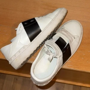 Valentino Open sneakers vit/svart - Snygga Valentino Open sneakers i vitt skinn med bred svart läderdetalj över snörningen. Klassisk rund tå och platt sula med ikoniska nitar längs kanten. Perfekt för dig som gillar stilrena och lyxiga sneakers med en cool twist.