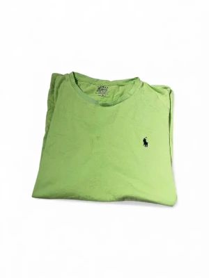 Grön t-shirt från Polo Ralph Lauren - Fräsch grön t-shirt från Polo Ralph Lauren med klassisk rund hals och korta ärmar. T-shirten har den ikoniska Polo-loggan broderad i mörkblått på bröstet. Tillverkad i mjuk bomull som är skön mot huden och passar perfekt till jeans eller shorts.