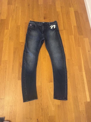Blå jeans från Hampton Republic 27 - Snygga blå jeans från Hampton Republic 27 med vit tryckt siffra '27' på fickan. Klassisk femficksmodell med röda sömmar och dragkedja. Jeansen har en rak passform och är tillverkade i slitstarkt denimtyg. Perfekt för dig som gillar stilrena detaljer. Storlek 158, inte mycket använda, perfekt skick