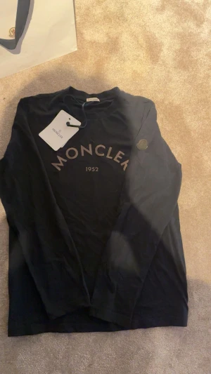 Svart långärmad Moncler t-shirt - Svart långärmad t-shirt från Moncler med stort tryck 'MONCLER 1952' på bröstet och en diskret logopatch på ärmen. Tillverkad i mjuk bomull som känns skön mot huden. Perfekt för dig som gillar stilrena och exklusiva plagg.