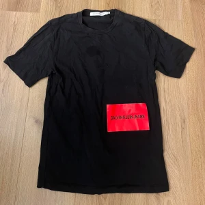 Svart t-shirt Calvin Klein  XS - Svart t-shirt från Calvin Klein i storlek XS. Framsidan har en stor röd patch med loggan och baksidan har en vit patch med loggan i svart. Klassisk rund hals och korta ärmar. Materialet är mjuk bomull, perfekt för en clean och modern look.