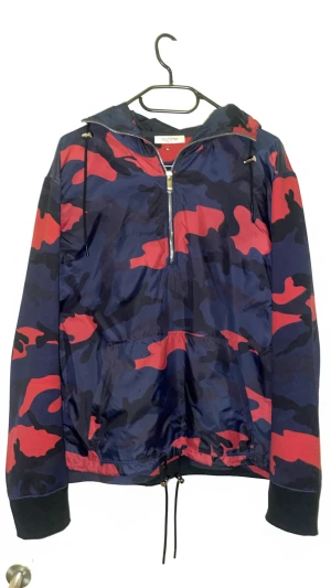 Valentino jacka - Snygg och extremt sällsynt vindjacka från Valentino med blå, mörkblå och röd camouflage-mönster. Jackan har huva med snörning, half zip-dragkedja och ribbade muddar. Perfekt för dig som vill sticka ut med en exklusiv look. Finns äktahetsbevis!