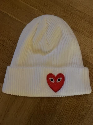 Vit ribbad mössa med hjärtdetalj - Säljer en vit ribbstickad mössa med ett broderat rött hjärta med ögon framtill. Mössan är mjuk och stretchig, perfekt för att hålla värmen och ge en clean look. Passar dig som gillar enkel stil med en twist.