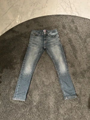 Tommy Jeans Justin Slim Tapered 29/30 - Säljer ett par blå jeans från Tommy Jeans, modell Justin Slim Tapered. Jeansen har en snygg tvätt med slitningar och klassiska fem fickor. De är smala i benen och har en modern, avsmalnande passform. Materialet är jeans med stretch för extra komfort. 