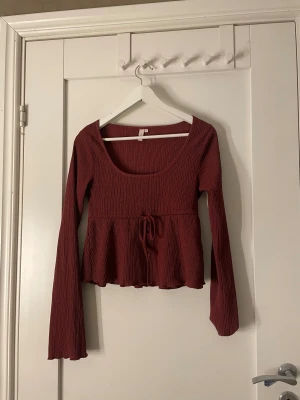 Vinröd långärmad topp från Nelly - Säljer en vinröd långärmad topp från Nelly i storlek S. Toppen har en rundad halsringning, ribbad struktur och en söt knytning i midjan. Modellen är lätt croppad med volangkant nedtill. 
