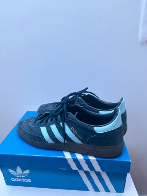 Adidas Spezial mörkblå sneakers mocka - Adidas Spezial sneakers i mörkblå mocka med ljusblå detaljer och klassiska tre ränder på sidan. Skorna har snörning, platt brun sula och rund tå. Märket syns i guld på sidan och insidan har Adidas-logga. Snygg retrostil och sköna att bära.