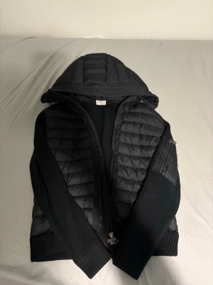 Svart cardigan från Moncler - Säljer en svart cardigan från Moncler med stickade partier och quiltad front. Västen har dragkedja, hög krage och ikonisk Moncler-logga på sidan. Perfekt för lager-på-lager och riktigt snygg till höst och vinter.