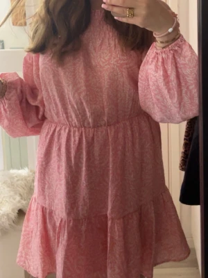 Rosa blommig kortklänning med volang - Supersöt rosa kortklänning med vitt blommönster och volangkant. Klänningen har långa puffärmar med resår vid handlederna och en avslappnad passform i midjan. Använd en gång under midsommar. Fler bilder finns skriv privat. 