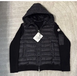 Svart Moncler pufferjacka med huva - Svart pufferjacka från Moncler med stickade ärmar och huva. Jackan har dragkedja framtill, ribbade muddar och en ficka med dragkedja på ärmen. Insidan har Moncler-etikett och serietidningsdetalj. Perfekt för dig som gillar exklusiv streetstyle.