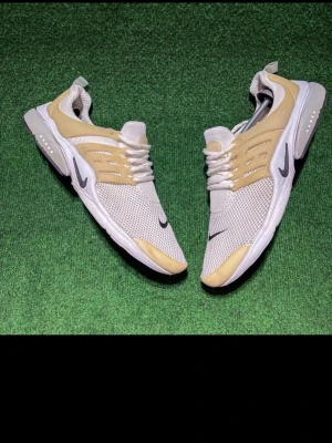 Nike Presto sneakers beige/vit - Nike Presto sneakers i beige och vit med mesh-ovandel och syntetiska detaljer. Snyggt svart swoosh på sidan och tå, samt platt sula och rund tå. Skorna har snörning och en flexibel passform som gör dem riktigt sköna för vardagsbruk. Perfekt för dig som gillar stilrena och sportiga sneakers.