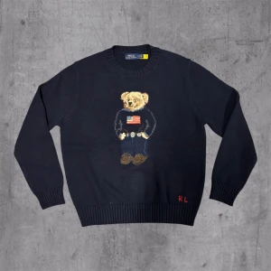 Mörkblå stickad tröja Polo Bear - Snygg mörkblå stickad tröja från Polo Ralph Lauren med klassisk Polo Bear broderad på bröstet. Tröjan har rund hals, ribbade muddar och RL-broderi vid nederkanten. Perfekt för dig som gillar ikoniska detaljer och stilren design.