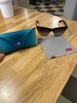 Svart Ray-Ban solglasögon med etui - Säljer ett par klassiska svarta Ray-Ban solglasögon med snygga mörktonade glas. Bågarna är mattsvarta och loggan syns både på glaset och sidan. Medföljer ett blått Ray-Ban etui och putsduk. Perfekt accessoar för soliga dagar!