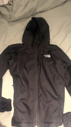 Svart vindjacka från The North Face - Svart vindjacka från The North Face i storlek M. Jackan har huva, hel dragkedja och meshfoder på insidan. Snygg logga på bröstet och minimalistisk design. Perfekt för blåsiga dagar och outdoor-vibes.