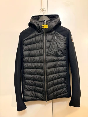 Svart pufferjacka från Parajumpers - Svart pufferjacka från Parajumpers med huva och coolt gult spänne vid halsen. Jackan har quiltad framsida och rygg, dragkedjor på fickor och bröst, samt ett snyggt PJS-märke på ärmen. Materialmix med polyester och syntet, perfekt för höst och vinter.