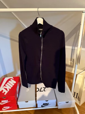 Svart kofta i merinoull från Brothers - Stilren svart kofta från Brothers i mjuk merinoull. Modellen har lång ärm, hög krage och hel dragkedja framtill. Perfekt för dig som gillar klassisk look med modern touch. Passar dig som vill ha en enkel men snygg tröja till garderoben. Krymt i tvät så passar S