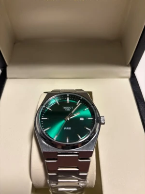 Grön Tissot PRX klocka med stållänk - Snygg Tissot PRX klocka med grön urtavla och stilren stållänk. Klockan har minimalistiska index, datumfunktion och klassisk rund boett. Perfekt accessoar för dig som gillar modern design och schweizisk kvalitet. Levereras i original ask.
