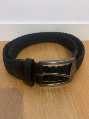 Svart flätad bälte - Snyggt svart flätat bälte med robust metallspänne och detaljer i konstläder. Bältet har en stilren look och passar perfekt till jeans eller chinos. Flätningen ger en cool textur och gör det enkelt att justera längden efter behov.