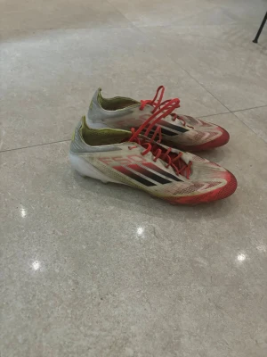 Adidas F50 fotbollsskor röd/vit - Säljer ett par Adidas F50 fotbollsskor med snygg design i vitt, rött och inslag av gult. Skorna har snörning och platt sula med fasta dobbar för gräsplan. Lätta och smidiga med svart Adidas-logga på sidan. Perfekt för dig som vill ha speed på planen.