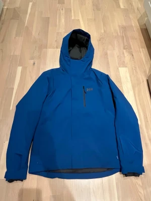 Skidjacka från Helly Hansen, S - Snygg blå skidjacka från Helly Hansen med huva, snölås, ficka på armen för liftkort, bröstficka med dragkedja, innerficka med dragkedja, sidofickor med dragkedja, fleece på insidan av dragkedjan som skydd för hakan, vädringsmöjlighet under armarna (med dragkedja), justerbara ärmslut. Mjuk och lätt jacka som är fodrad, vind och vattentät. Mycket fint skick, inga tecken på användning. Regular fit, storlek S. Inköpt 2024 för 1800 kr. 