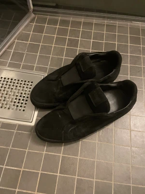 Svarta sneakers från Axel Arigato - Svarta sneakers från Axel Arigato med stilren design och bred kardborreband över foten. Skorna har en rund tå och är tillverkade i mocka med insida i skinn. Platt sula och diskret branding på innersulan. Perfekt för dig som gillar minimalistisk stil. Bra skick för super pris! Kontakta vid frågor.