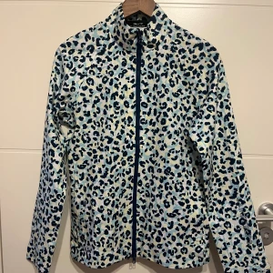 Leopardmönstrad regnjacka J.Lindeberg - Snygg regnjacka från J.Lindeberg med blått och vitt leopardmönster. Jackan har hög krage, hel dragkedja och lång ärm. Perfekt för dig som vill sticka ut även när det regnar. Materialet är lätt och syntetiskt för att stå emot väta.