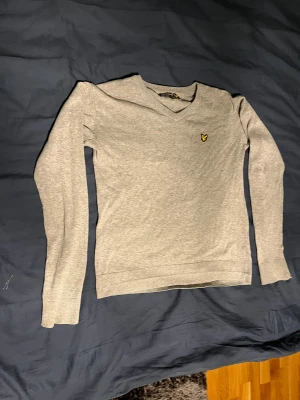 Grå v-ringad tröja Lyle & Scott - Grå v-ringad tröja från Lyle & Scott med deras klassiska gula fågel-logga på bröstet. Tröjan har långärmade ribbade muddar och är tillverkad i mjuk bomull. Perfekt för dig som gillar stilrena och enkla plagg med diskret branding.