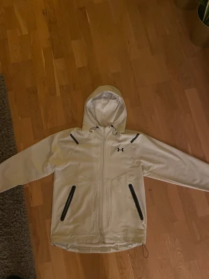 Beige vindjacka från Under Armour - Snygg Beige/Vit vindjacka från Under Armour med svart logga på bröstet. Jackan har huva med snörning, svarta dragkedjor på fickorna och hel dragkedja framtill. Materialet är syntet och insidan har mesh för extra ventilation. Perfekt för dig som gillar sportig stil. Endast använda ett par gånger 